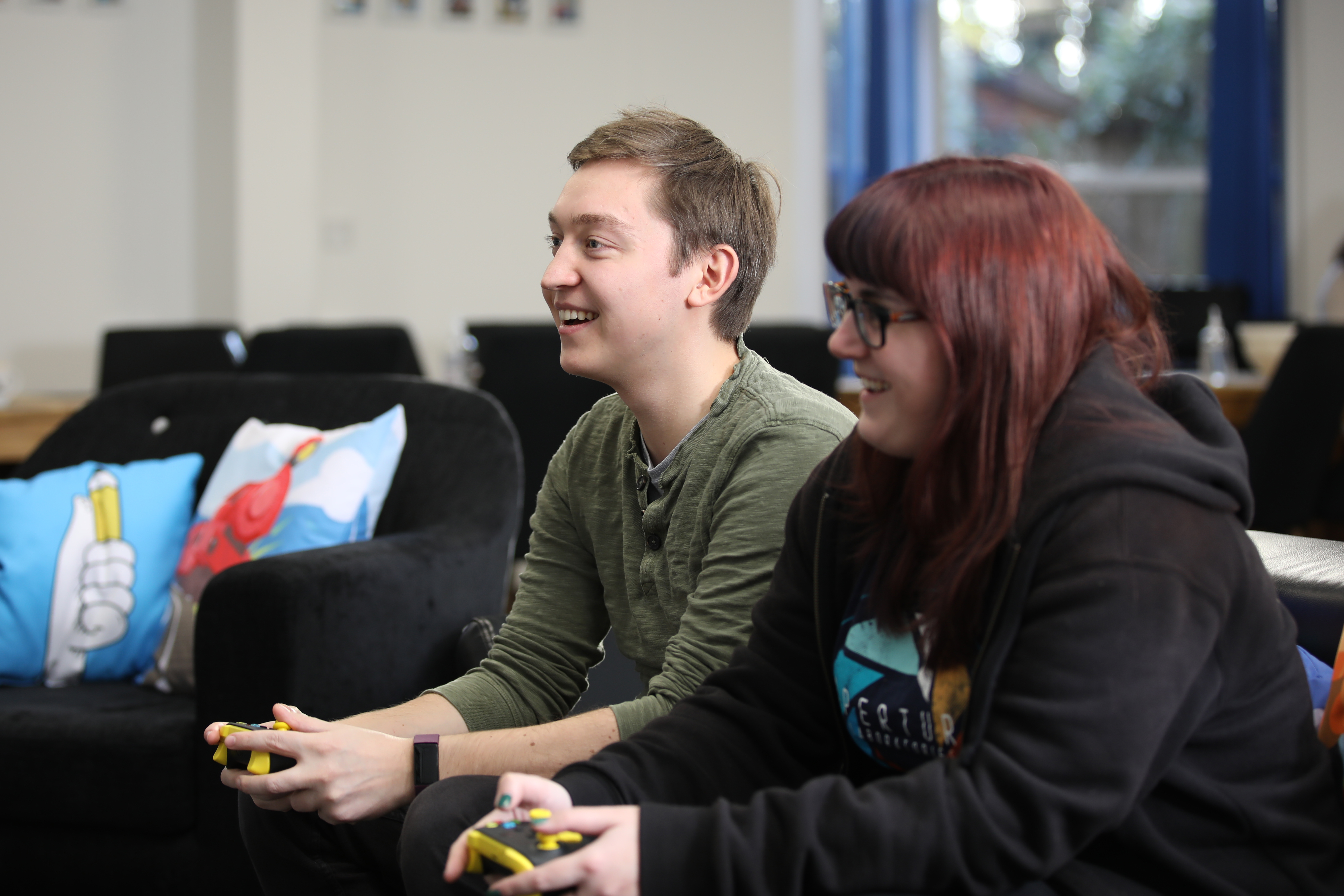 Image shows two members of the kwalee team looking at a TV holding play controllers. One is wearing a black hoody, red hair and glasses. The other has a green tshirt. They are both smiling at the game they are playing. 