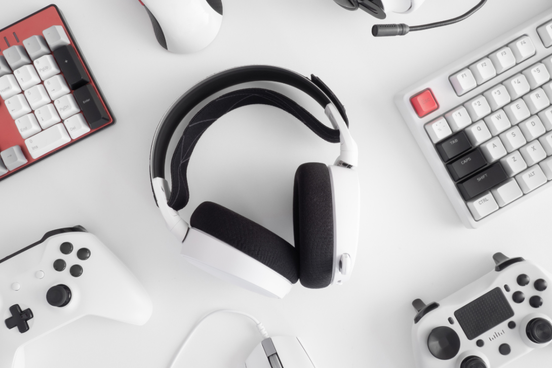 An assortment of modern technology accessories including headphones, keyboards, and PlayStation controllers neatly arranged on a clean white background, suggesting a setup for immersive gaming or audio entertainment.