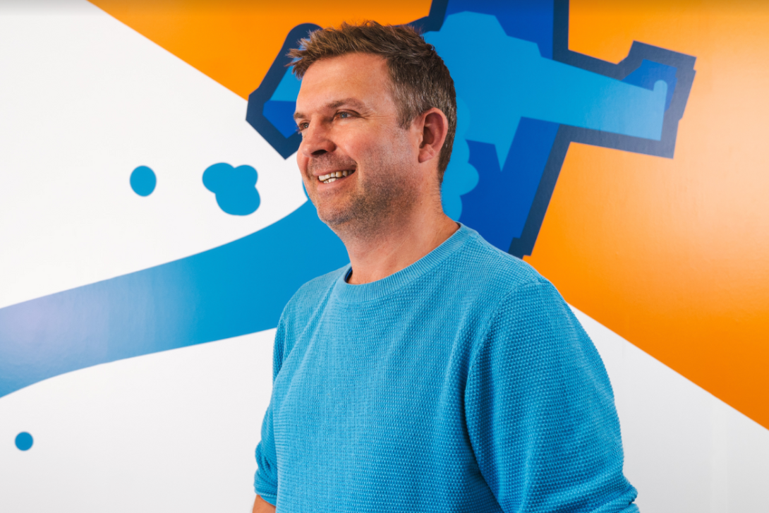 Harry, representing his blog Maverick Marketing, smiles confidently in a light blue jumper against a backdrop of vibrant orange, white, and blue shapes. His positive demeanor reflects the innovative spirit of Maverick Marketing strategies.