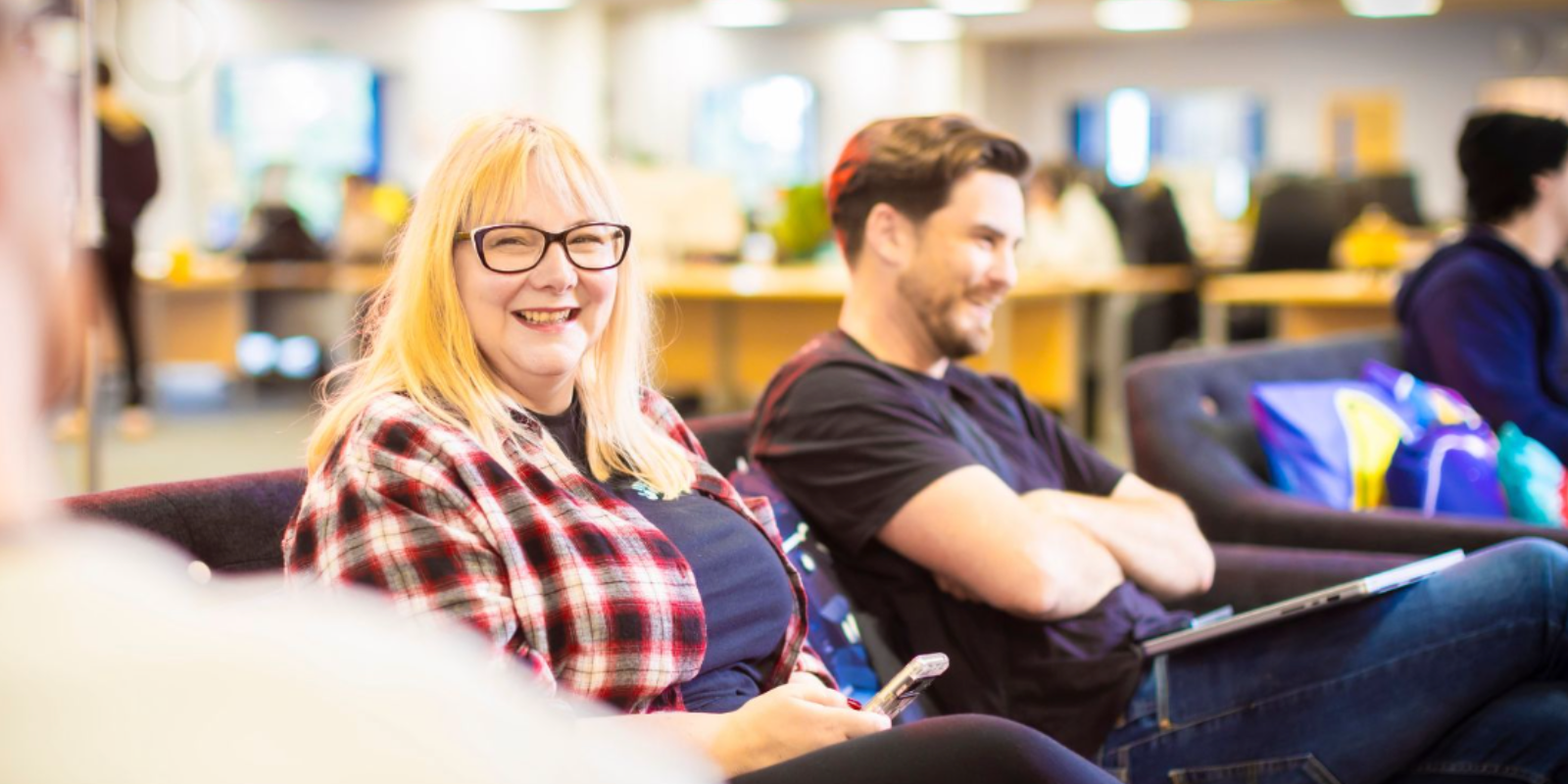 A diverse group of people sitting in the Kwalee lounge. Jen is facing the camera sitting down, she has blonde hair and a block fringe.  She is wearing a red chequered flannel shirt with a navy top underneath. She is sat next to Tom who is head od development here at Kwalee. Jen is looking at the camera smiling. 