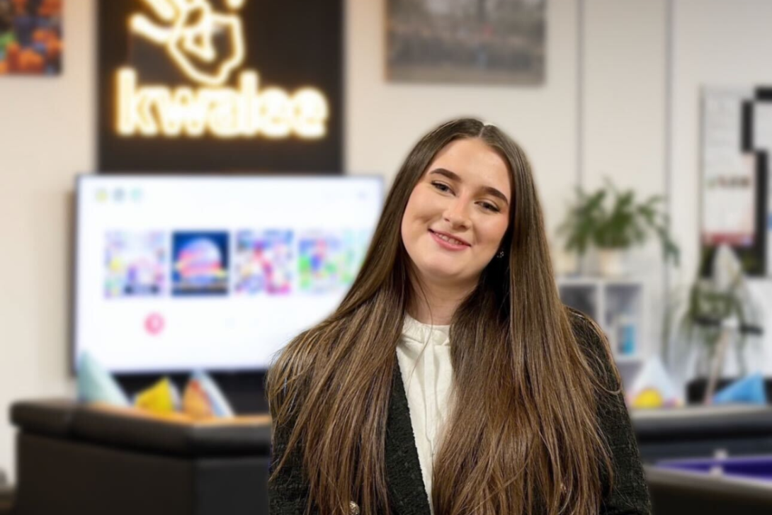 Lizzy, our apprentice in administration, stands in Kwalee's vibrant office space, featuring a TV, pool table, and the iconic yellow Kwalee logo light. She wears a black blazer, embodying dedication to her apprenticeship in games.