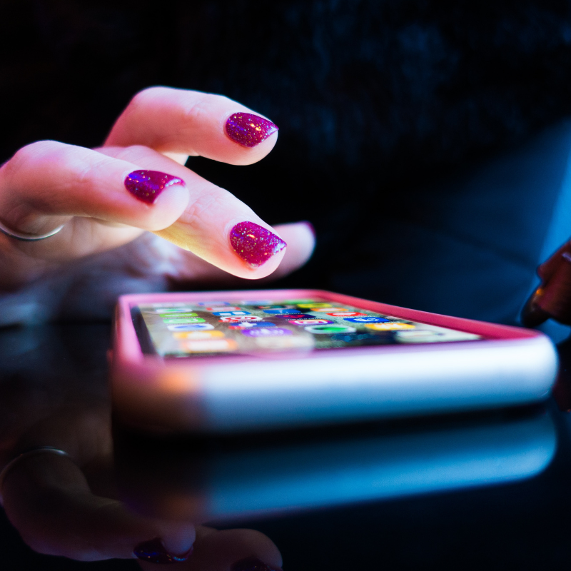 Image shows a very close up photo of a mobile phone on a flat surface. There is a hand above it, looking like it is swiping thriough the phone looking for the correct app. They have pink nail varnish on, covered in glitter, and also a silver shiny ring. 