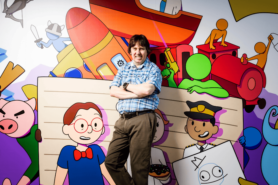 David Darling, smiling and standing in front of a wall painted with characters from Kwalee's games