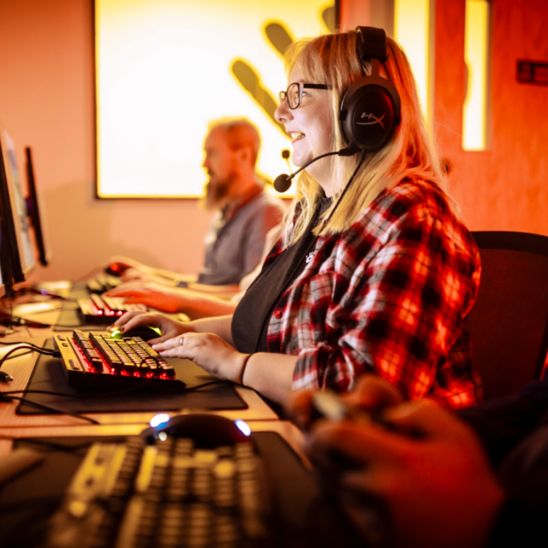 Image shows Jen in a room with red lighting, facing a computer playing a video game. She is wearing a headset with a microphone, like she is playing online with s group of friends. William Cox is in the background, playing on the computer to, but he is slightly faded. 